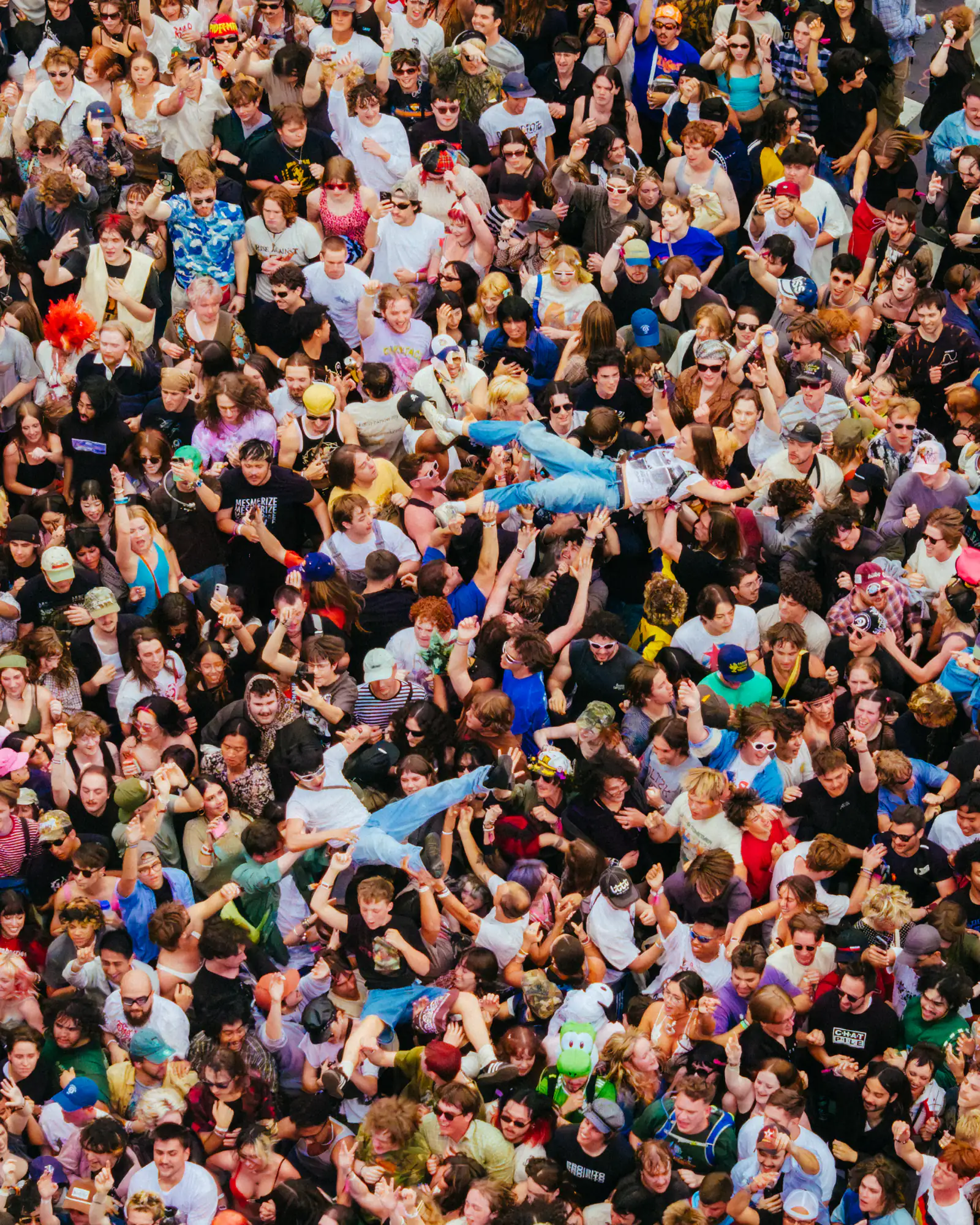 A packed festival crowd seen from above, arms up, with two people crowd surfing across it